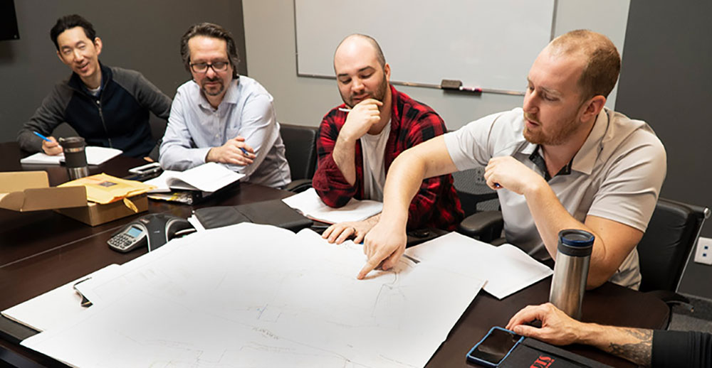Team of engineers collaborating in a meeting room, reviewing detailed network planning blueprints on a table, with one team member pointing to a specific section of the design.